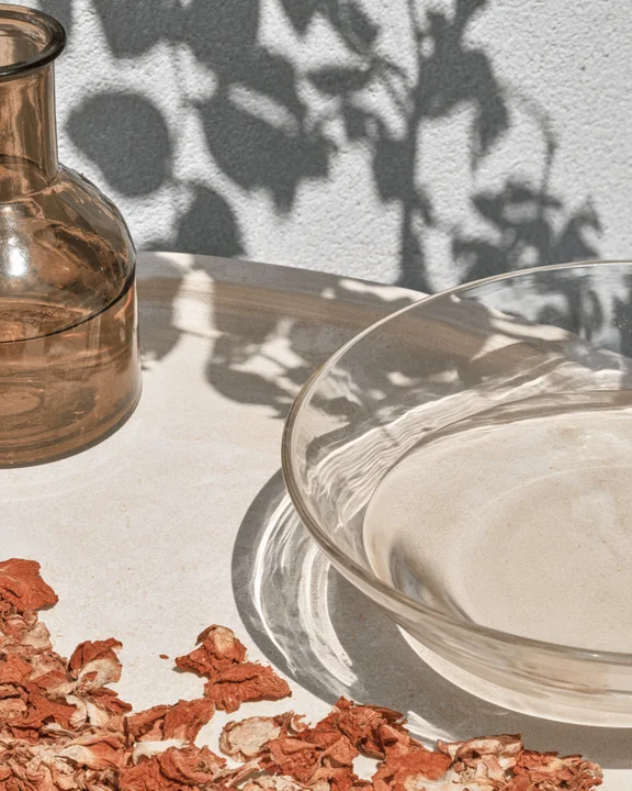 Small amber glass bottle and a shallow ceramic bowl of rose water on a pale plaster surface with dried petals