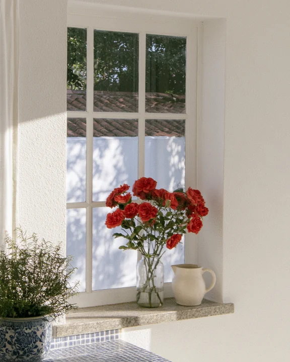 Kitchen window sill with a jar of short-cut roses, herbs and a ceramic jug