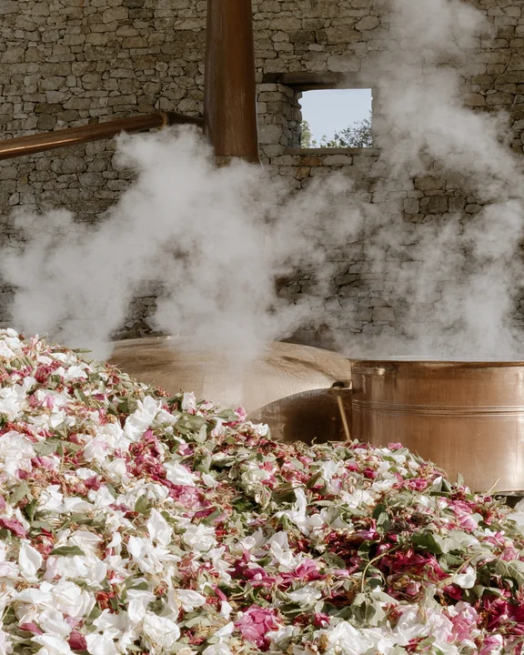 Traditional copper still in a stone-walled rose distillery with heaped fresh petals beside it and steam in the morning light