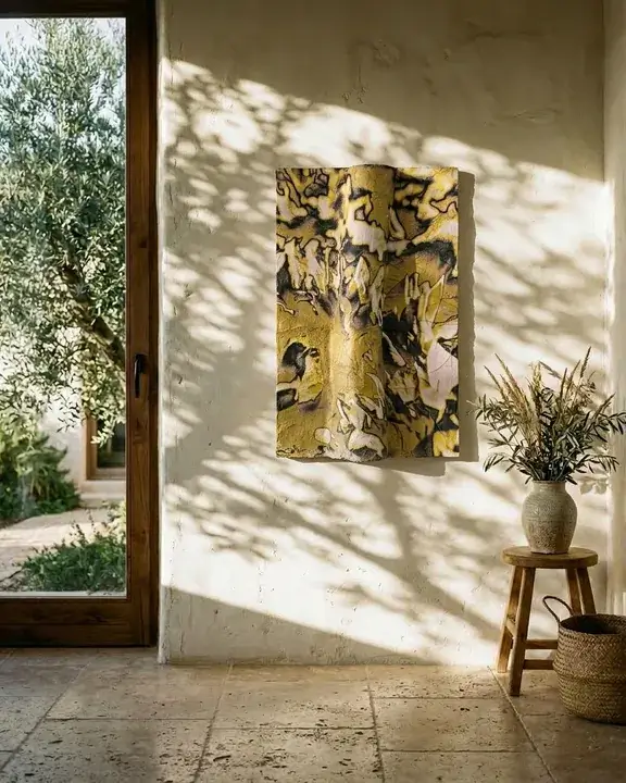 Dappled shadow from an olive tree falling across a lime-wash wall and across a COBELIO relief hung on it, its ochre surface catching the late afternoon light