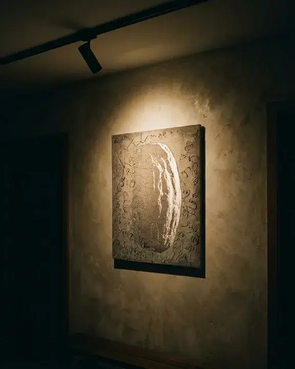 A ceiling track spot aimed down at a COBELIO pumpkin relief on a plaster wall, its warm beam grazing the surface while the rest of the passage stays dark