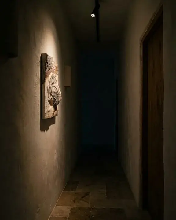 A narrow plaster hallway at dusk, a COBELIO relief on the left wall picked out by a small ceiling spot, the far end of the corridor in deep shadow